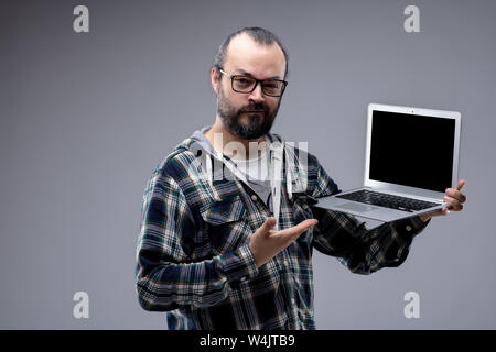 Gerne Mann, der auf seinem Handheld Laptop mit einem Lächeln Nachweis angezeigt oder die Ausstellung einer Bestätigung, leerer Bildschirm mit Kopie Raum über Grau Stockfoto