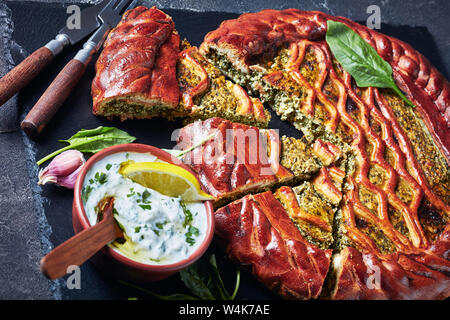 In Scheiben geschnittenen Spinat Ricotta Torte auf einem schwarzen Schiefer Fach mit Tzatziki Sauce in einer Schüssel auf einer konkreten Tabelle, Ansicht von oben, close-up Stockfoto