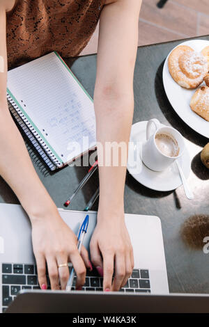 Blick von oben auf die Frauen Hände schreiben auf einem Laptop - Espresso Kaffee, Brötchen und ein Notebook auf dem Tisch - Bild vertikal Stockfoto