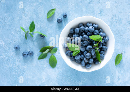 Blaubeeren mit Blätter, weiße Schüssel auf blauem Hintergrund, Ansicht von oben Stockfoto