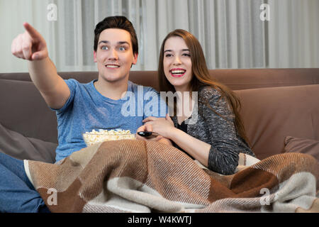 Junges Paar beobachten Film auf dem Sofa gut aussehender Mann Halteplatte mit Popcorn und etwas auf dem TV-Bildschirm schöne Frau und umarmte ihn Stockfoto