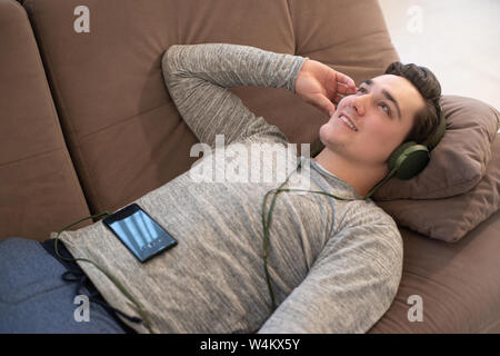 Jungen gutaussehenden Mann lächelnd auf dem Sofa hören die Musik im Kopfhörer vom Smartphone Gefühl entspannt Stockfoto