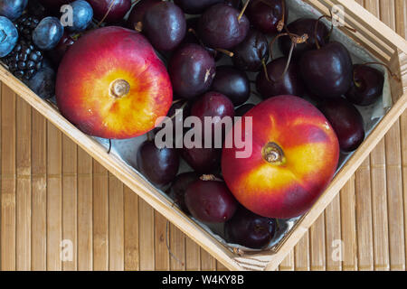 Stilleben mit Früchte. Himbeeren, Kirschen, Blaubeeren, Brombeeren und Nektarine. In einer kleinen Kiste auf einer Matte. Stockfoto