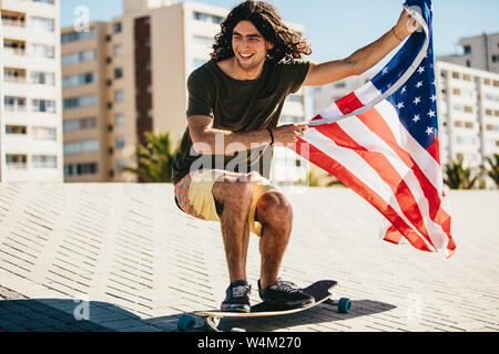 Glückliche junge Mann skateboarding mit USA-Flagge im Freien in der Stadt. Mann mit der amerikanischen Flagge auf dem Skateboard. Stockfoto