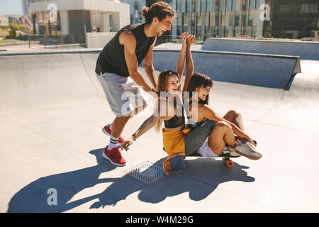 Gruppe von Jugendlichen Spaß mit Skateboard in den Skate Park. Lächelnd Kerl drücken zwei Mädchen auf einem Skateboard im Freien. Stockfoto