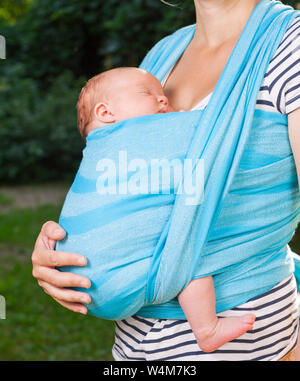 Junge Mutter ihr kleines Baby girl in blue Schlinge im Freien. Kind schläft in Komfort, Sicherheit und Schutz. Stockfoto