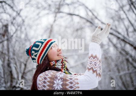 Foto der lächelnde Mädchen auf gestrickte Mütze und Schal fallende Schneeflocken im Winter Wald bei Tag Stockfoto