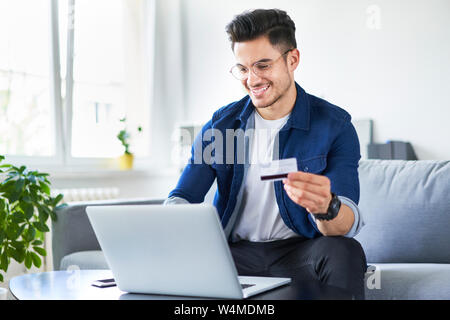 Portrait von gutaussehenden jungen Mann zahlen Sie online mit Kreditkarte und Laptop zu Hause sitzen Stockfoto