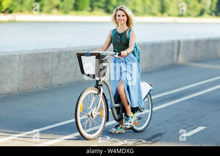 Bild des blonden Jungen in langen Jeans Rock sitzen auf dem Fahrrad auf der Straße in der Stadt Stockfoto