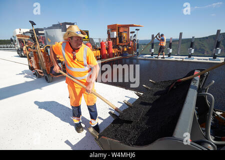 Zeltingen Rachtig, Deutschland. 24. Juli, 2019. Arbeitnehmer Asphalt der Straße Oberfläche des Hochmosel Brücke im Bau bei Temperaturen deutlich über 30 Grad Celsius. Es gibt keine schattigen Platz auf dem 160 Meter hohen Gebäude über dem Moseltal. Quelle: Thomas Frey/dpa/Alamy leben Nachrichten Stockfoto