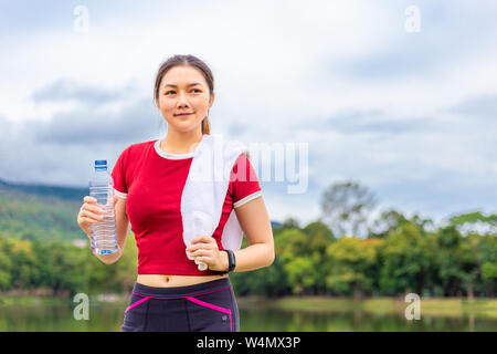 Schöne asiatische Athleten Frau trinkt Wasser während ihrer Pause vom Morgen Übung an einem See Park mit Blick auf den See und die Berge im Hintergrund, Stockfoto