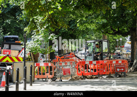 Sicherheitsbarrieren surround Männer mit einem kleinen Bagger in Cheltenham Town Centre auszugraben Stockfoto