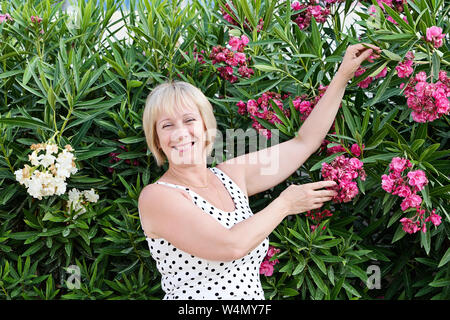 Kaukasische Dame mittleren Alters stellt sich und lacht fröhlich in blühenden Blumen der Oleander. Körper positive und Wechseljahre Konzept, ehrliche Portrait Stockfoto