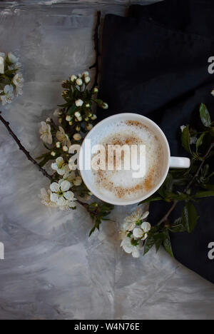 Blick von oben auf die weiße verführerischen Blumen neben einer warmen Tasse Cappuccino auf schwarz Serviette auf graue Oberfläche. Stockfoto