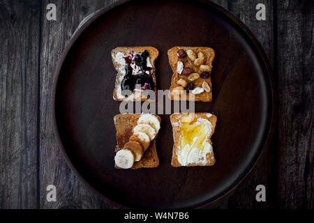 Blick von oben auf die verschiedenen Toppings auf Toastbrot, Erdnussbutter, Banane, Marmelade, Honig auf Holz- Hintergrund. Stockfoto