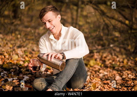 Foto eines Mannes gießt Tee aus einer Thermoskanne in eine Tasse in den Wald Stockfoto