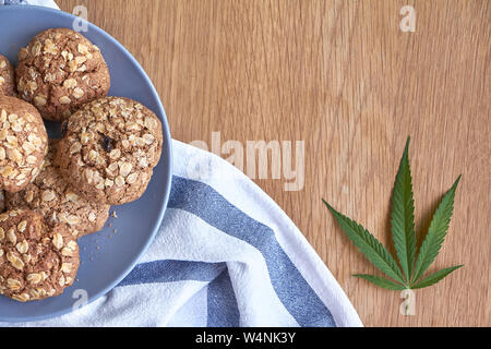 Ansicht von oben ein paar Haferflocken Topf Cookies auf graue Platte auf ein gestreiftes Handtuch auf hellem Holz- Oberfläche und einem Blatt von Marihuana neben, selektiver Fokus Stockfoto