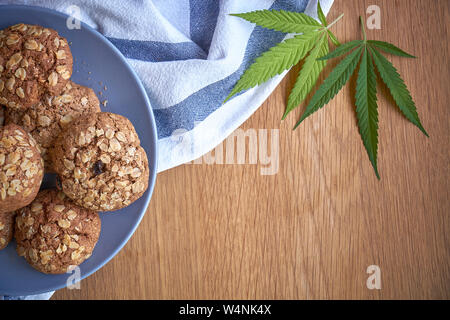 Ansicht von oben ein paar Haferflocken Topf Cookies auf graue Platte auf ein gestreiftes Handtuch auf hellem Holz- Oberfläche und zwei Blätter von Marihuana neben, selektiver Fokus Stockfoto