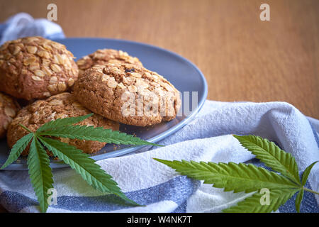 Nahaufnahme von ein paar Haferflocken Topf Cookies auf eine graue Platte auf ein gestreiftes Handtuch auf hellem Holz- Oberfläche und zwei Blätter von Marihuana neben, selektiver Fokus Stockfoto