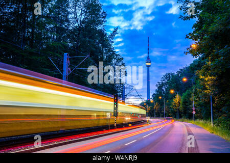 Deutschland, beleuchtete Straßenbahn vorbei Straße der Verkehr neben dem Fernsehturm Stuttgart Stadt im grünen Wald Natur Landschaft in magischen twili Stockfoto