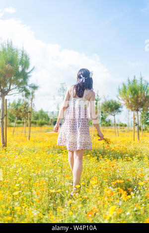 Ansicht der Rückseite eine schöne Frau in Kleid zu Fuß durch ein Feld von Blumen ihre Schuhe tragen