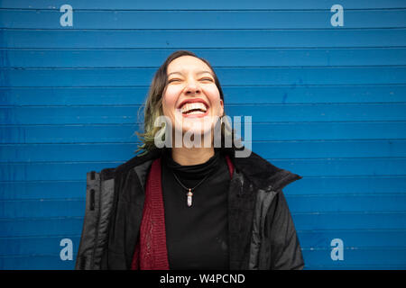 Lachende junge Frau mit geschlossenen Augen auf der blauen Wand, im Regen. Stockfoto