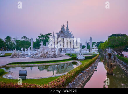 Die abendlichen Spaziergang um den Weißen Tempel (Wat Rong Khun) mit Blick auf den schönen Ubosot, Brücke der Wiedergeburt Zyklus, Teich, formschnitt Garten, Wassergraben mit Stockfoto