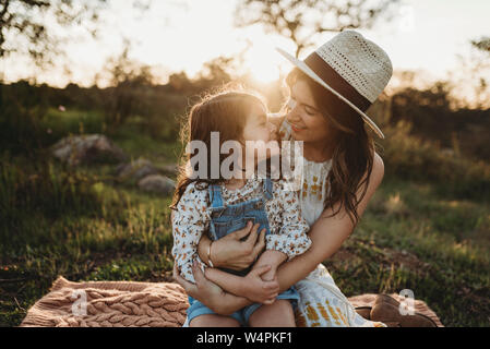 Mutter und Tochter sitzen auf Decke in Wiese mit Hintergrundbeleuchtung Stockfoto