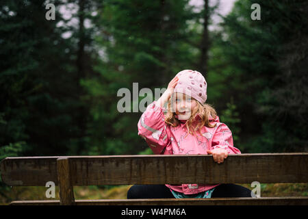 Adorable kleine Mädchen in rosa Jacke sitzt hinter Holzzaun und Einstellung beanie auf Hintergrund mit unscharfen dunklen Wald in der Nähe von Reykjavik. Stockfoto