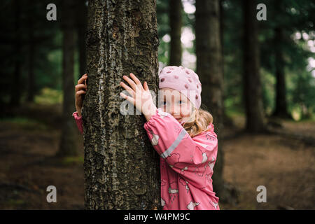 Süße kleine blonde Mädchen mit rosa Regenmantel und Hut lächelnd und umarmt Baum in Reykjavik, Island Wald weg schauen Stockfoto