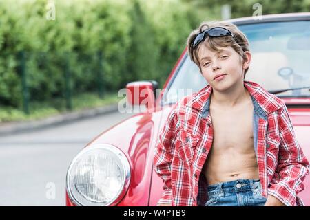 Junge, 10 Jahre alt, mit Sonnenbrille und Plaid Shirt stützt sich auf ein rotes Auto und sieht cool aus in die Kamera, Deutschland Stockfoto