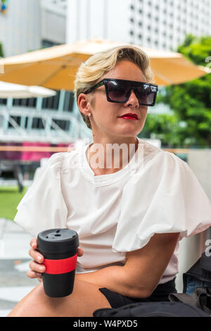Business Frau mit Sonnenbrille trinken Kaffee aus Schwarz Rot wiederverwendbare Schale Garten Singapur konzentriert Stockfoto
