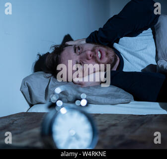 Schlaflose und verzweifelten jungen kaukasischen Mann wach nachts nicht schlafen zu können, frustriert und beunruhigt, schaut auf die Uhr leiden unter Schlaflosigkeit. Stockfoto