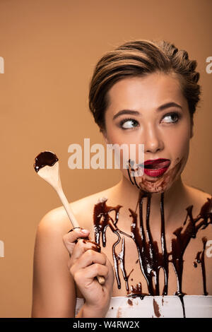 Schöne Frau lecken roten Lippen, holding Holzlöffel und Wegsehen mit Schokolade Flecken auf der Haut isoliert auf beige Stockfoto