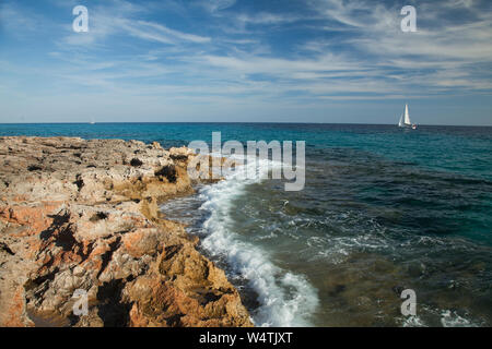 Blick vom felsigen Ufer auf hellen, weißen Yacht segeln auf dem Mittelmeer blau leuchtenden Meer an einem sonnigen Tag mit einer weißen Wolken am blauen Himmel. Stockfoto