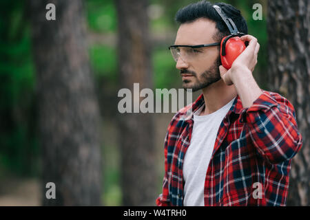 Nachdenklich Holzfäller berühren Noise Cancelling Kopfhörer und Wegsehen im Wald Stockfoto