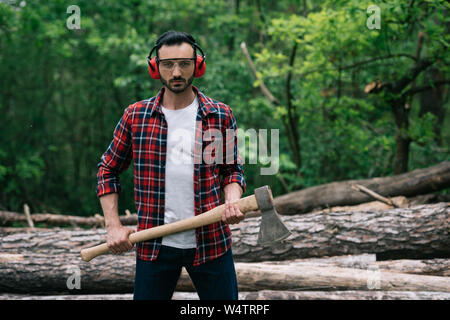 Ernsthafte Holzfäller in Schutzbrille und Noise Cancelling Kopfhörer holding Ax und Kamera Stockfoto