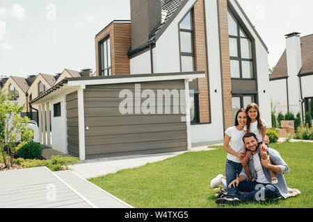 Familie sitzt auf Rasen im Hinterhof, großes, modernes Haus auf Hintergrund Stockfoto