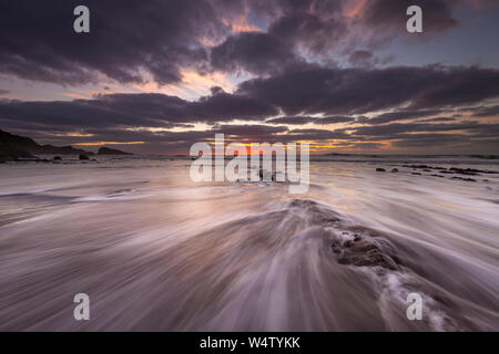Wellen, Strömung über den Sand und Felsen bei Sonnenuntergang am Welcombe Mund Beach in North Devon, UK. Stockfoto