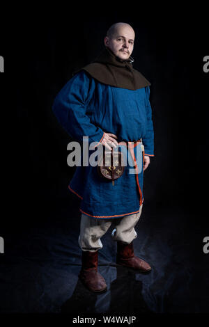 Ein glatzköpfiger Mann verkleidet in legere Kleidung der Wikingerzeit (Hut, Kaftan, Hosen, Haube) steht auf einem schwarzen Hintergrund. Stockfoto