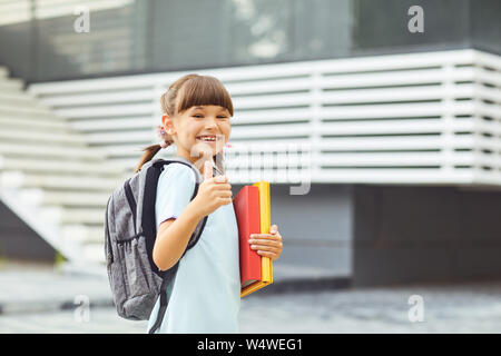 Lächelnde Mädchen Schülerin mit einem Rucksack und Bücher in der Hand geht in die Schule. Schüler Mädchen, dass Daumen hoch, beim Stehen auf der Straße. Stockfoto