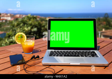 Laptop mit grünen Bildschirm geöffnet auf Holztisch mit dem Smartphone und dem angeschlossenen Kopfhörer und das blaue Meer Panorama im Hintergrund in selectiv Stockfoto