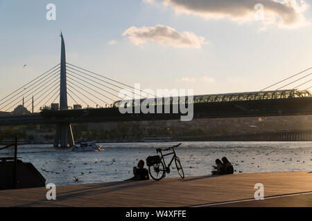 Istanbul, Türkei - 19 Juli, 2019: ein Biker, sein Fahrrad und zwei junge Frauen sind an der Küste des Goldenen Horns auf Holzboden in Sonnenuntergang. Stockfoto