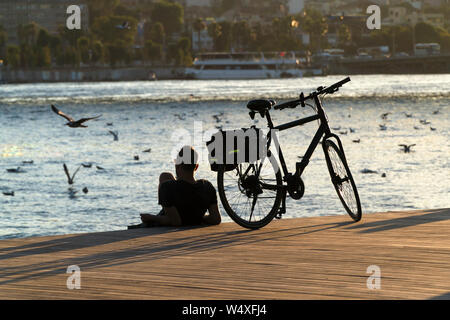 Istanbul, Türkei - 19 Juli 2019: ein Biker und sein Fahrrad liegen an der Küste des Goldenen Horns auf Holzboden in Sonnenuntergang. Stockfoto