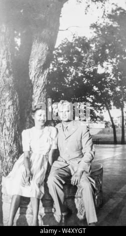 Junge Frau in einem Kleid, setzt sich unter einen Baum mit ihrem Ehemann, in militärischer Uniform gekleidet, beide lächeln, San Francisco, Kalifornien, 1943. () Stockfoto