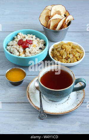 Frühstück: Haferflocken mit Obst Stücke und Kandierte Erdbeeren, goldenen Rosinen, Apfel Chips, Honig in kleinen Schüsseln und eine Tasse schwarzen Tee auf einem grauen Holz Stockfoto