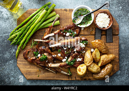 Gegrillte Lammkoteletts mit grünen Kräutersoße, Feta, Spargel und Kartoffeln auf einem Holzbrett serviert. Stockfoto