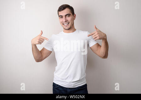 Portrait von gutaussehenden jungen Mann mit perfekten weißen Lächeln in Weiß leer t-shirt zeigt mit seinen Fingern, Platz zum Design Stockfoto