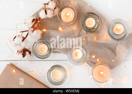 Schöne brennende Kerzen mit Baumwolle Blumen und Girlanden auf weißem Holz- Hintergrund Stockfoto