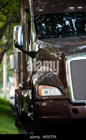 Klassische glänzend Big Rig Motorhaube dunkelbraun Semi Truck Schlepper steht vor einer anderen semi Trucks auf den Parkplatz auf der Straße mit grünen Bäumen ein Stockfoto